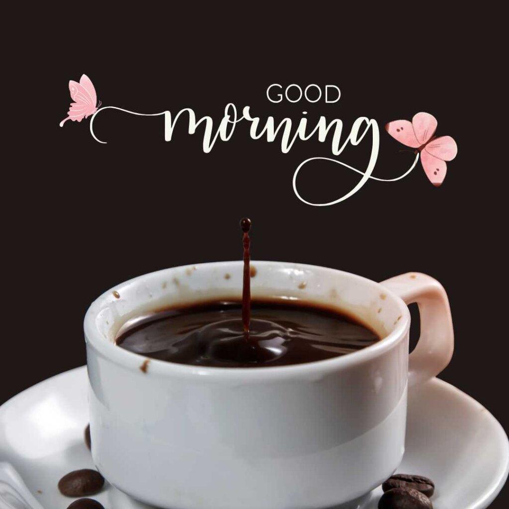 A close-up shot of a single drop of dark coffee splashing upward into a white cup, sitting on a saucer with scattered coffee beans. The background is a solid dark brown with an elegant cursive 'Good Morning' text greeting decorated with pink butterflies.