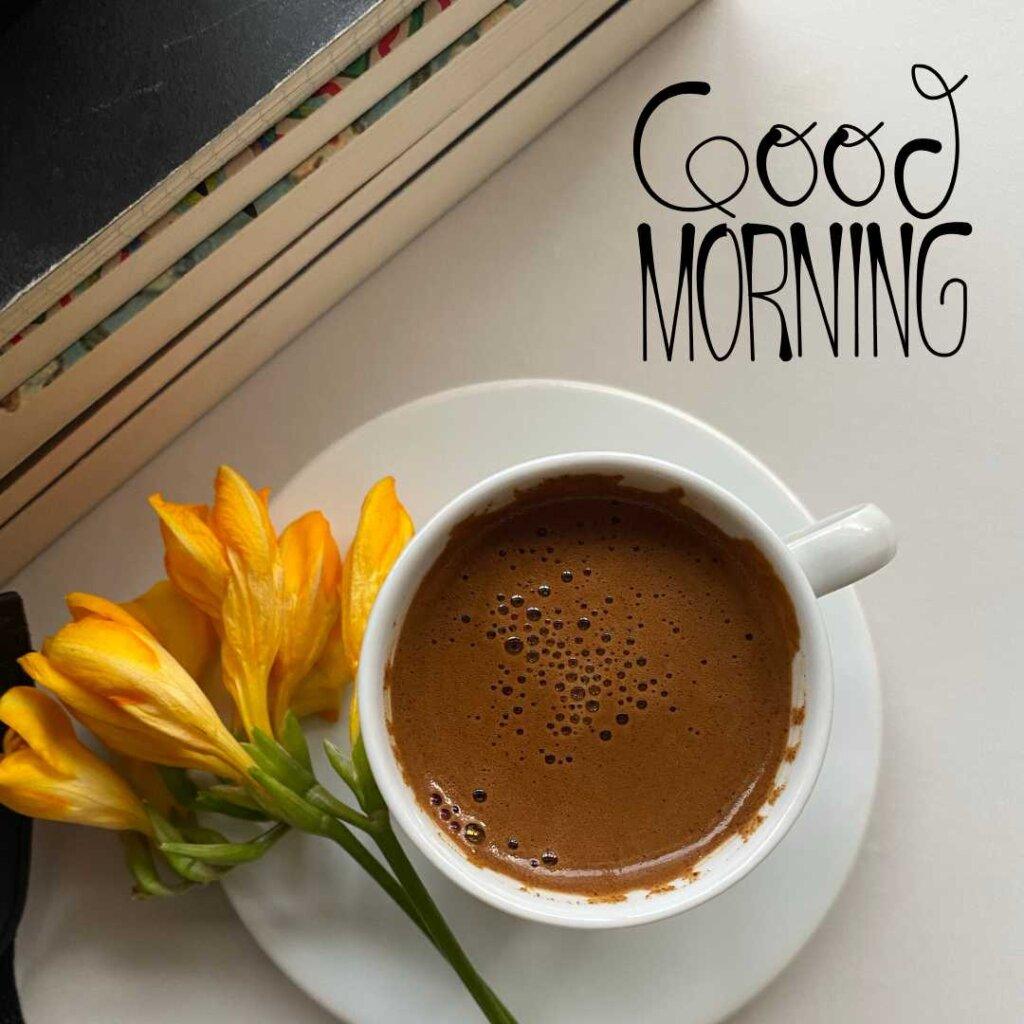A cup of hot coffee with rich froth on a white saucer, placed next to bright yellow flowers and stacked books with a 'Good Morning' text overlay.