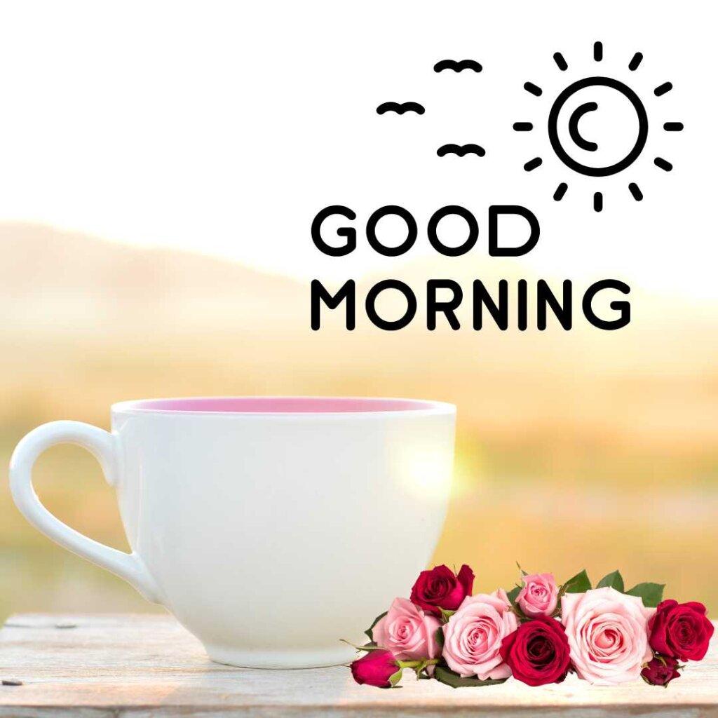 A large white tea cup on a rustic wooden surface next to a small bouquet of pink and red roses, set against a blurred golden sunrise background with 'Good Morning' text and minimalist sun and birds illustrations.