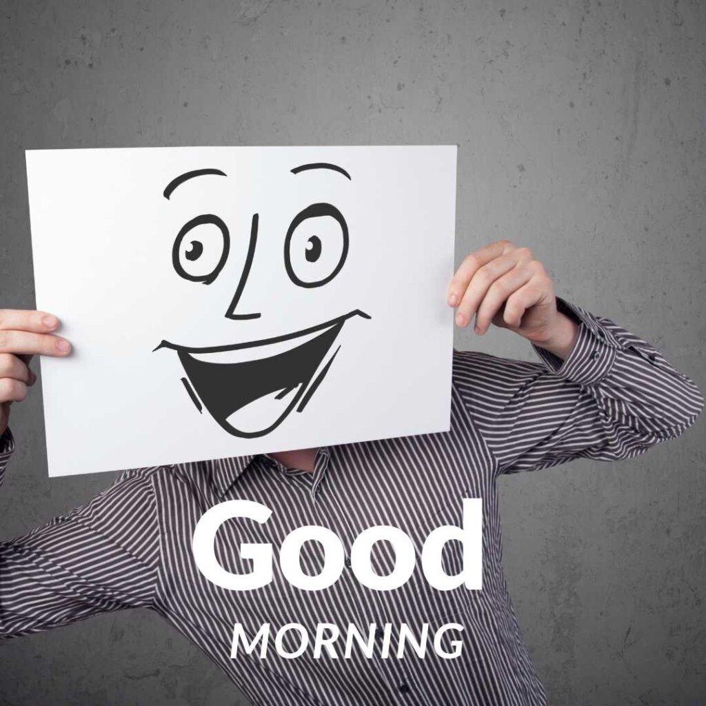 A person hiding their face behind a white sign with a funny wide-eyed happy doodle, featuring bold "Good Morning" text against a concrete background.