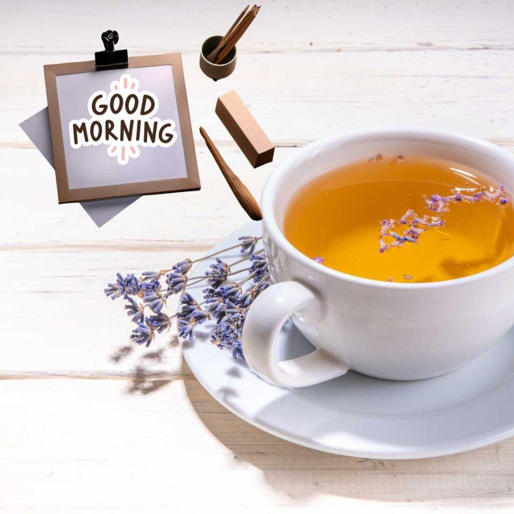 A white ceramic cup filled with golden herbal tea and floating lavender petals, sitting on a saucer with dried lavender stems on a white wooden table. A small clip-on frame features a 'Good Morning' text greeting.
