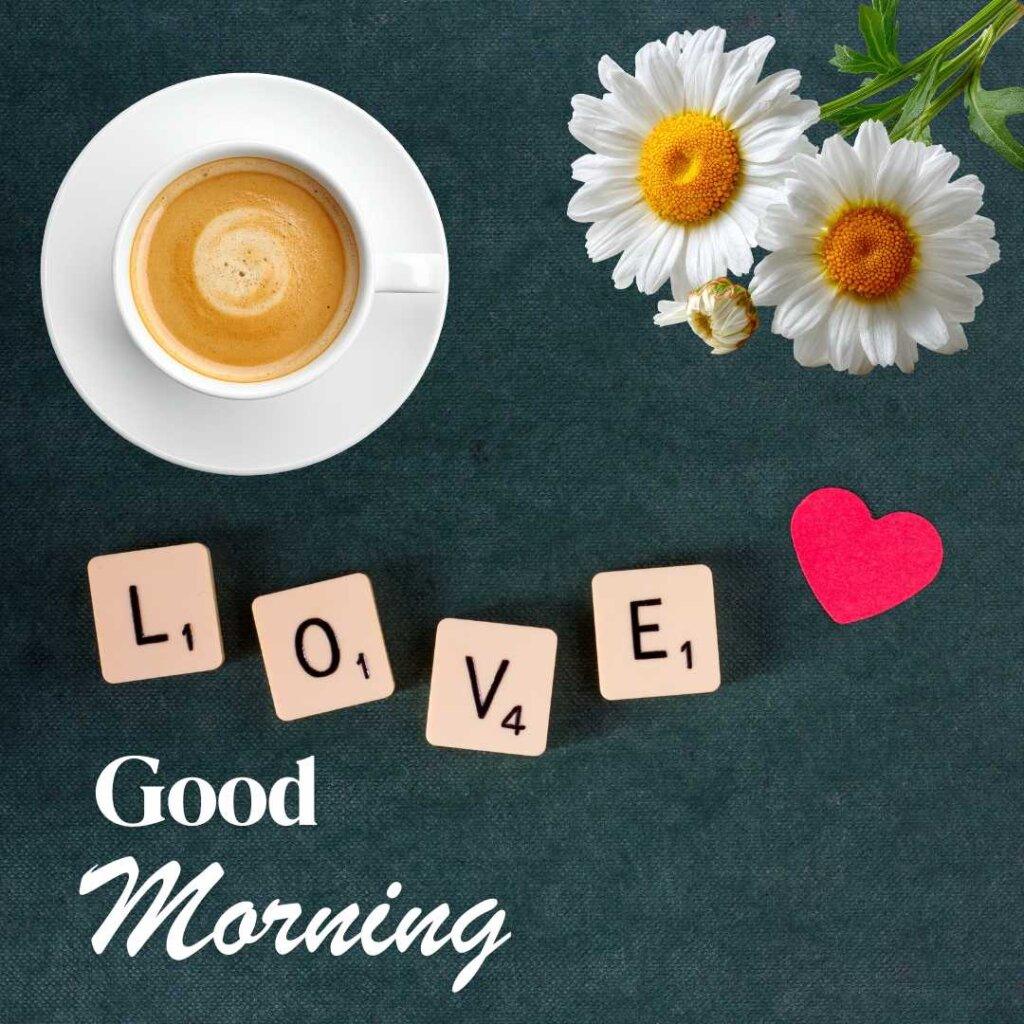 A top-down view on a dark textured background featuring a white cup of coffee with creamy froth, two white daisy flowers, a small red paper heart, and scrabble tiles spelling out 'LOVE' above a white 'Good Morning' text greeting."