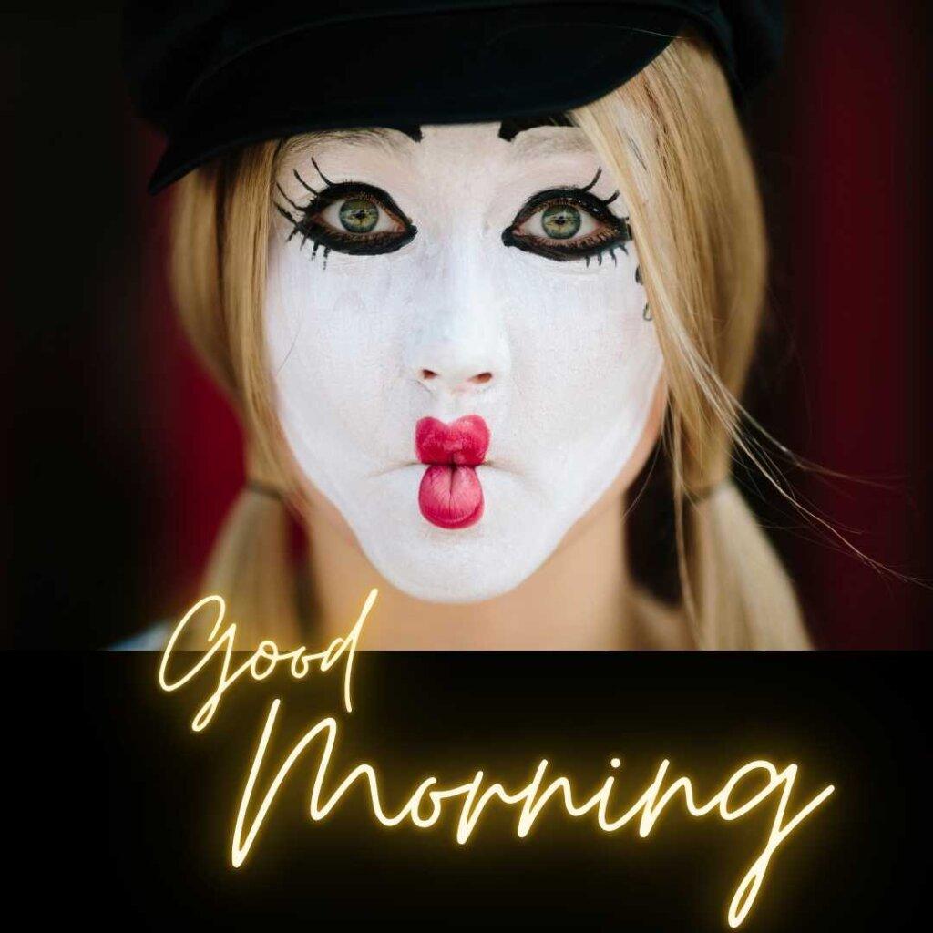 Close-up of a female mime artist with white face paint, dramatic eye makeup, and red lips making a kissing expression, with glowing yellow "Good Morning" neon script.