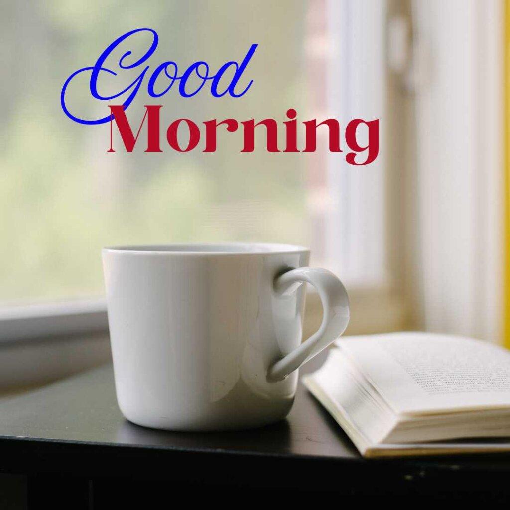 A white ceramic mug sitting next to an open book on a dark table by a bright window, with a 'Good Morning' text overlay in stylized blue and red fonts.