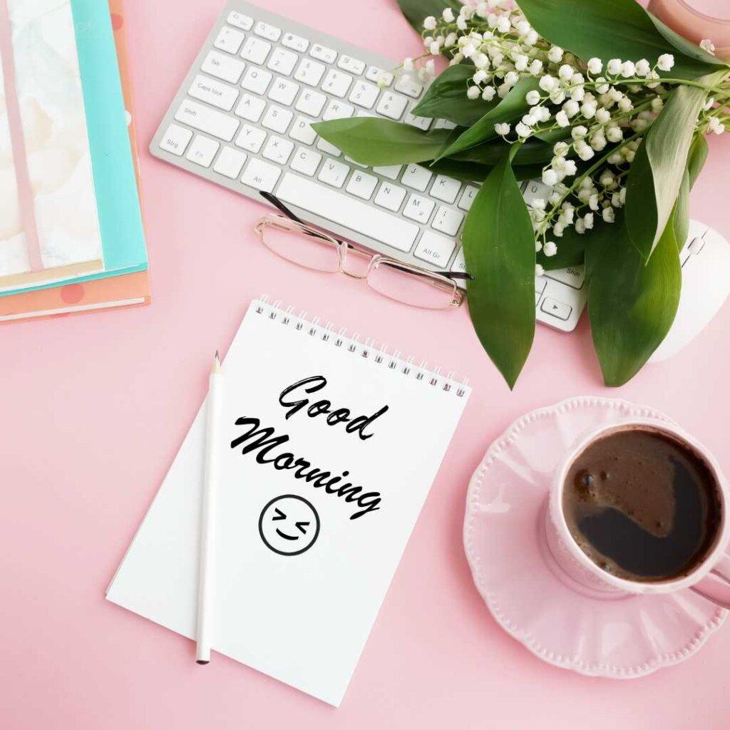 A top-down view of a pink desk setup featuring a white computer keyboard, reading glasses, a bouquet of white lily of the valley flowers, a pink cup of black coffee, and a spiral notepad with 'Good Morning' text and a smiling emoji.
