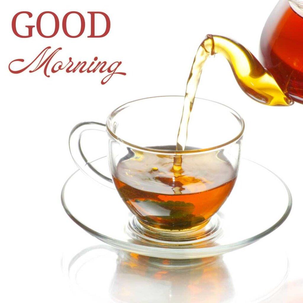 Fresh hot tea being poured from a glass teapot into a clear glass cup and saucer on a reflective white surface, with a red 'Good Morning' text greeting in the top left corner.