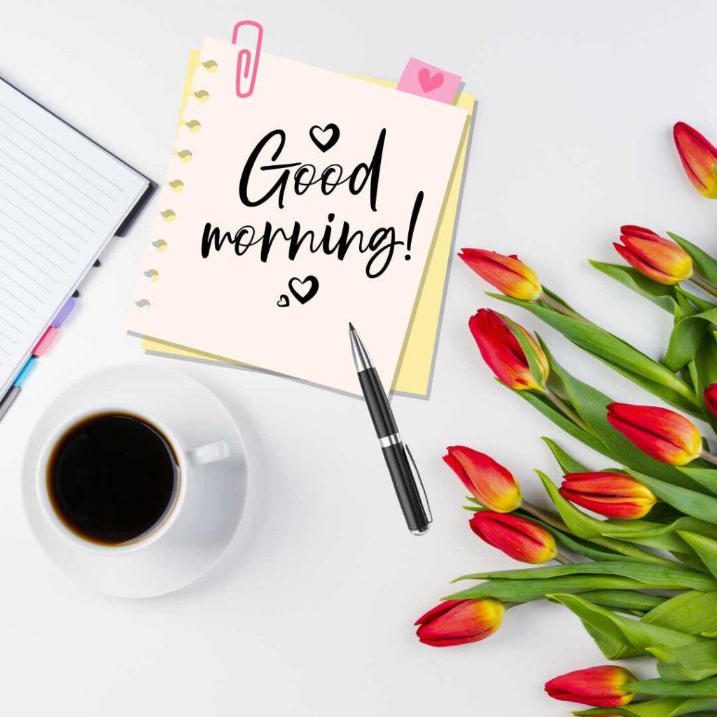 A flat lay composition on a white background featuring a bouquet of fresh red and yellow tulips, a white cup of black coffee, a sleek pen, and a sticky note with handwritten 'Good morning!' text decorated with small hearts.