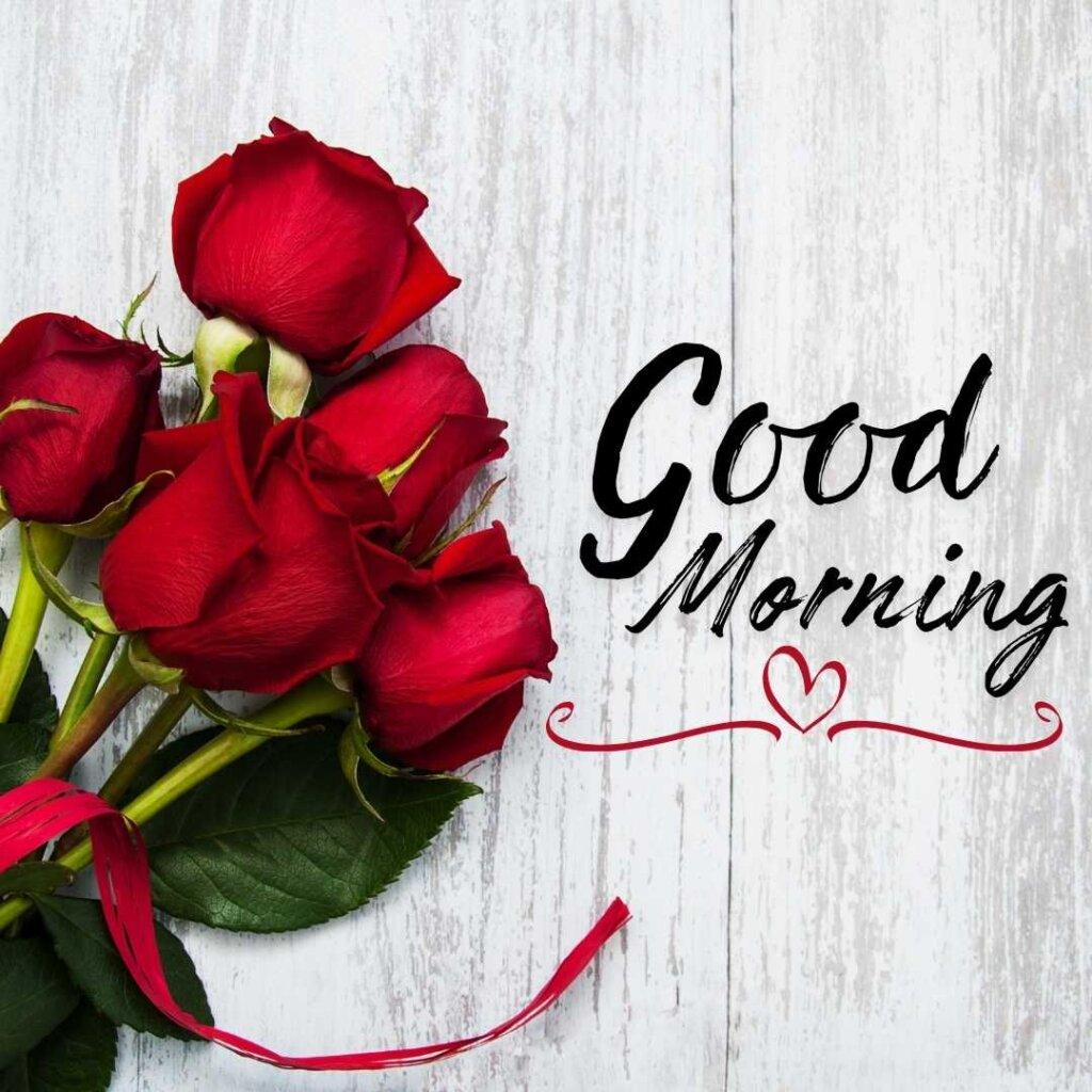 A beautiful bouquet of four fresh red roses tied with a red satin ribbon resting on the left side of a white rustic wooden plank floor, next to a stylish black cursive greeting Good Morning with a small red heart icon.
